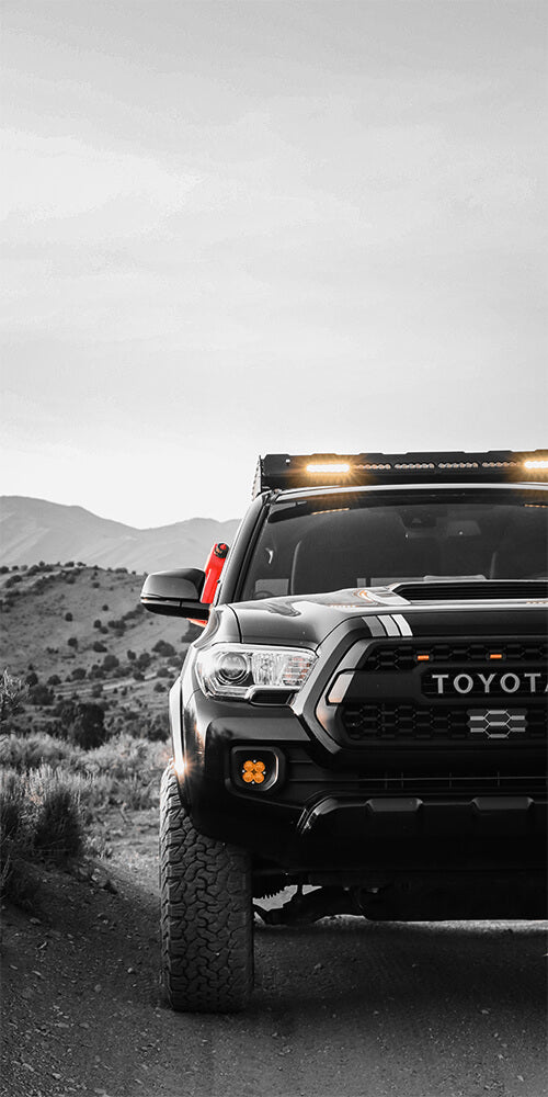 A rugged black Toyota truck with illuminated headlights and roof lights parked on a dirt trail.